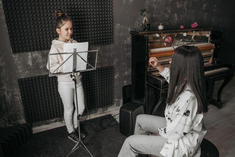 A young girl receiving vocal training from a woman near a piano in a music studio.